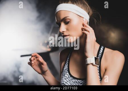 Selektive Fokussierung der Sportlerin mit Smartwatch berühren drahtlose Kopfhörer in Fitnessraum Stockfoto