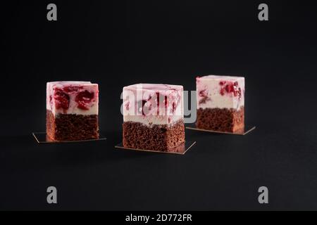 Nahaufnahme von drei leckeren frischen Scheiben Kuchen mit Keks und Mousse Schicht mit rotem Gelee und Kirschen isoliert auf schwarzem Hintergrund gemacht. Leckeres süßes braunes und rosafarbenes Dessert im Restaurant. Stockfoto