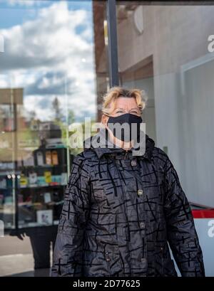 Eine ältere Frau in einer schwarzen Jacke und einer schwarzen medizinischen Maske geht aus der Tür eines Einkaufszentrums. Stockfoto