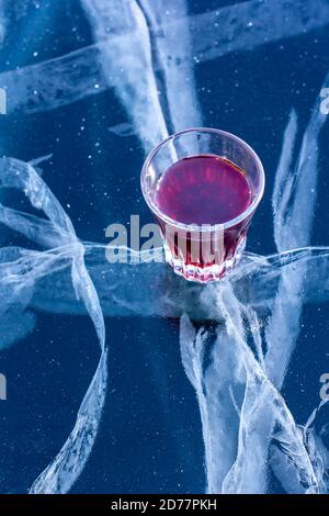 Ein Glas mit roter Tinktur steht auf dem Eis des Sees. Alkohol im Glas und Eis mit schönen tiefen Rissen. Vertikal. Stockfoto