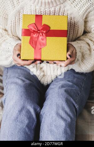 Junge Frau sitzt auf dem Bett in kuschelig gestrickten Wollpullover und hält Geschenkbox mit Rotgold. Hygge, Neujahr, Weihnachten, Weihnachtsvorbereitungen. Kand Stockfoto