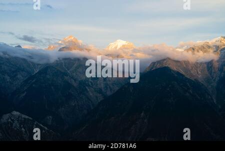 Schneebedeckte Berge unter Wolken und blauem Himmel und von Bäumen gesäumten Hängen, von erhöhter Position aus gesehen, bei Sonnenuntergang in Kalpa, Himachal Pradesh, Indien. Stockfoto