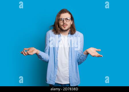 Verwirrt kaukasischen Mann mit Bart und lange Haare posiert Unentschlossen während der Geste mit Handflächen an einer blauen Studiowand Stockfoto