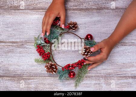 Damenhänden machen einen Weihnachtskranz aus Tannenzweigen, Zapfen und roten Beeren auf Holzbrettern Stockfoto