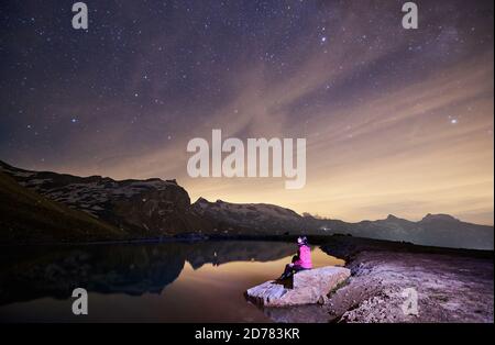 Landschaft in der Nacht, Frau auf Stein in der Nähe des Sees sitzen, schöne Berge mit Schnee auf dem Hintergrund. Wunderschöner Bergrücken mit hohen felsigen Gipfeln unter strahlendem Sternenhimmel. Konzept des Reisens in den Alpen Stockfoto