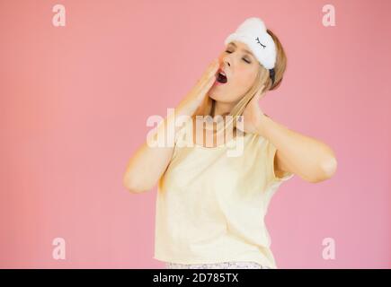 Müde junge Frau in gelben Schlafanzug zu Hause tragen Schlafmaske Gähnend, der Mund mit der Hand bedeckt, die zu Hause isoliert auf liegt Pastell rosa Hintergrund Studio po Stockfoto