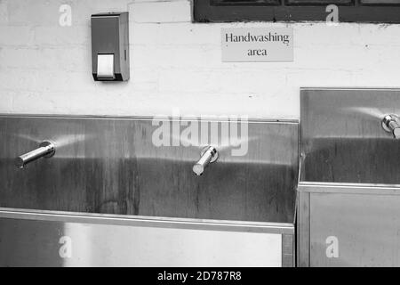 Nahaufnahme von Handwaschanlagen, Waschbecken aus Edelstahl und Wasserhähnen mit einem Seifenspender aus Edelstahl. Eine weiße Backsteinmauer im Hintergrund Stockfoto