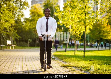 Glücklicher afroamerikanischer Geschäftsmann ist Roller im Park am sonnigen Tag reiten. Stockfoto