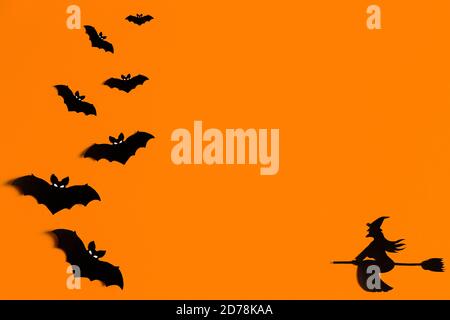 Silhouetten einer Fledermausschar und einer Hexe, die auf einem Besen aus schwarzem Papier auf orangefarbenem Hintergrund fliegt. Halloween Grußvorlage mit Kopierraum Stockfoto