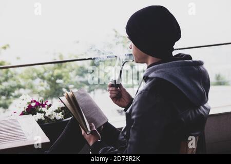 Rückansicht auf jungen schwarzhaarigen Mann, der in einem Café im Freien ein Buch durchbohrt und Tabakpfeife raucht. Stockfoto