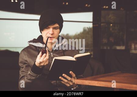 Rückansicht auf jungen schwarzhaarigen Mann, der in einem Café im Freien ein Buch durchbohrt und Tabakpfeife raucht. Braun getöntes Foto. Stockfoto