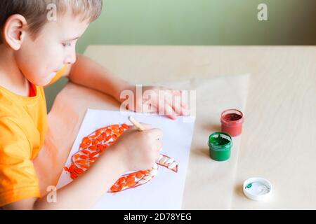 Schuljunge Kürbis aus Samen auf weißem Papier, Halloween DIY-Konzept. Schritt-für-Schritt-Anleitung. Schritt 3 Kürbiskerne mit oranger Farbe bemalen Stockfoto