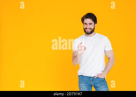 Ein glücklicher bärtiger junger Mann in einem weißen T-Shirt Und blaue Denim-Hosen lächeln an der Kamera und zeigen Geste OK Körpersprache alles wird in Ordnung sein auf einem Stockfoto