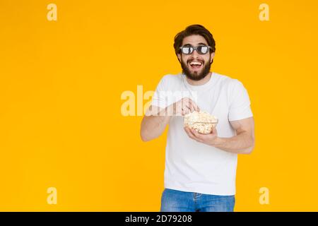 Bild einer lachen, glücklich, junger Mann in lockeren weißen T-Shirt essen Sie Popcorn mit 3D-Brille ansehen Film über dem gelben Hintergrund isoliert. Stockfoto