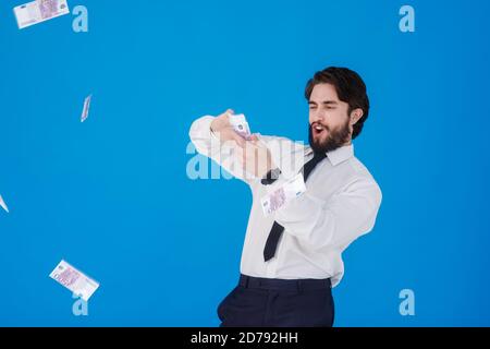Ein junger fröhlicher bärtiger Geschäftsmann in einem weißen Hemd und schwarzer Krawatte auf blauem Hintergrund spritzt Geld. Verrückter Kerl wirft Geldscheine. Studio Stockfoto
