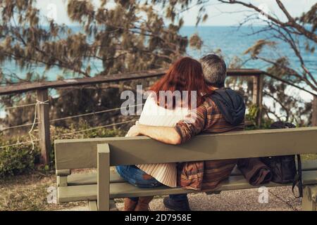 Glückliches Paar mittleren Alters verliebt sitzen auf einer Bank im Park mit Blick auf das Meer, umarmen und Blick auf das Meer. Stockfoto