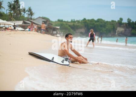 Der Kerl ruht sich auf einem tropischen Sandstrand aus, nachdem er eine Brandung gefahren ist. Gesunde aktive Lebensweise im Sommer Berufung. Stockfoto