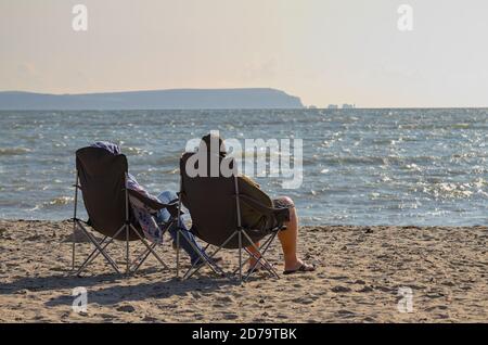 Mann Und Frau Packten Sich Gegen Die Kälte Und Genossen Den Sonnenschein Am Avon Beach UK mit Isle of Wight Needles in Die Entfernung Stockfoto
