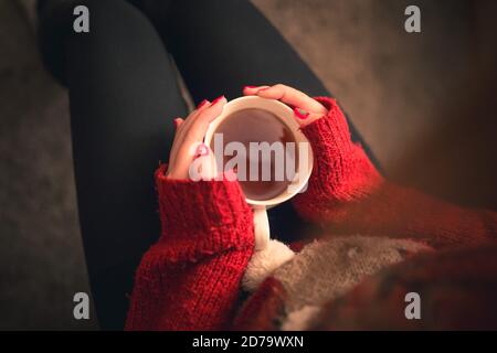 Top Down Aufnahme eines Mädchens mit einem wärmenden Teetrink in den Händen auf dem Sofa sitzend mit weihnachtlich gefärbten Fingernägeln und Rentierpullover. Stockfoto