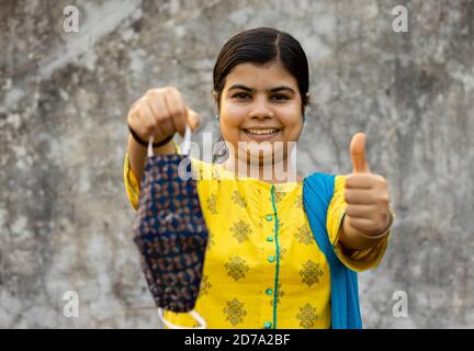 Indische Frau in gelbem Kleid hält hausgemachte Baumwolle Maske und Daumen nach oben als ähnliche Geste Stockfoto