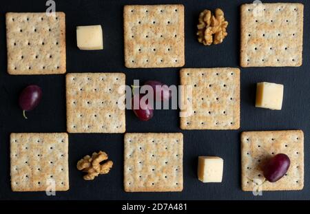 Maasdam Käse, Walnüsse, Kekse und lila Trauben vor einem dunklen Hintergrund gestaffelt. Poster. Stockfoto