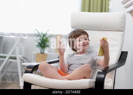 Happy Kid Junge sitzen auf dem Computer mit Laptop und mit einem Videoanruf. Lächelndes Kind sitzt zu Hause Rest auf Stuhl beschäftigt mit Gadgets. Online-Bildung und Stockfoto