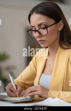 Fokussierte Geschäftsfrau in Brillen tragen gelbe Strickjacke macht eine Notiz in Notebook. Office weibliche Manager aus der Ferne arbeitet von zu Hause während der zweiten w Stockfoto