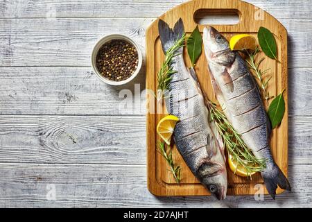 Seebarsch frischer Fisch entkaltet und entkalkt auf einem Schneidebrett mit frischem Rosmarin, Zitronenkeilen und Lorbeerblatt auf einem Schneidebrett auf einem hölzernen Hintergrund, t Stockfoto