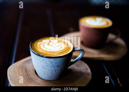 Zwei Tassen heißen Cappuccino auf Holztisch mit Löffeln auf dunklem Tischhintergrund. Frühstückszeit. Geschlossen. Stockfoto