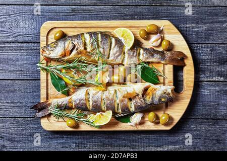 Gebratener Fisch mit Zitronenkeilen und Kräutern: Rosmarin, Petersilie und Lorbeerblatt, serviert auf einem Holzbrett mit grünen Oliven und Tartar Stockfoto