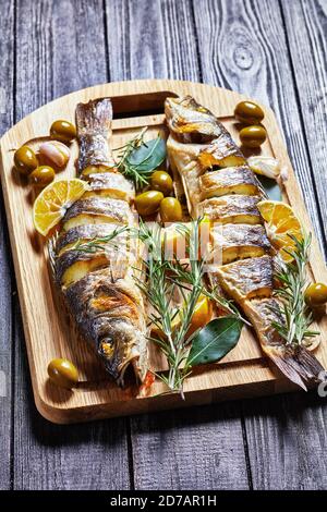 Gebratener Fisch mit Zitronenkeilen und Kräutern: Rosmarin, Petersilie und Lorbeerblatt, serviert auf einem Holzbrett mit grünen Oliven und Tartar Stockfoto