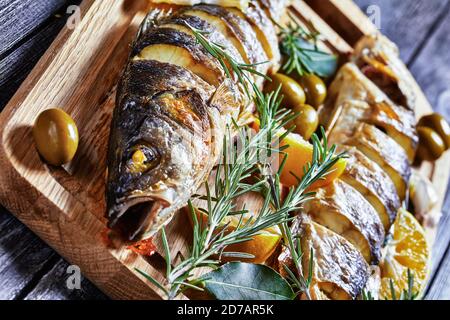 Gebratener Fisch mit Zitronenkeilen und Kräutern: Rosmarin, Petersilie und Lorbeerblatt, serviert auf einem Holzbrett mit grünen Oliven und Tartar Stockfoto