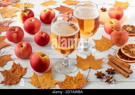 Reife frische saftige rote Äpfel, Gewürze und Gläser mit Apfelwein auf einer hellen Holzoberfläche mit Herbstblättern. Horizontale Ausrichtung, selektiver Fokus. Stockfoto