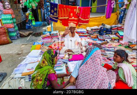 Freundlich lächelnder Stallholder sitzt an einem Stall, der bunte Stoffe in einem Straßenmarkt in einem Dorf in Madhya Pradesh, Indien verkauft Stockfoto