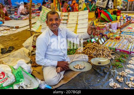 Stallholder sitzt in einem Stall, der lokale Produkte verkauft, die Ingwer auf Waage wiegen, auf einem Straßenmarkt in einem Dorf in Madhya Pradesh, Indien Stockfoto