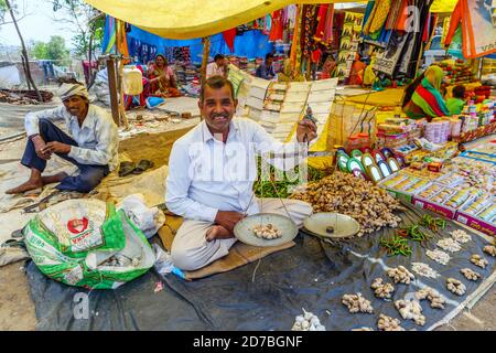 Stallholder sitzt in einem Stall, der lokale Produkte verkauft, die Ingwer auf Waage wiegen, auf einem Straßenmarkt in einem Dorf in Madhya Pradesh, Indien Stockfoto