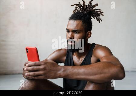 Bild von jungen afroamerikanischen Sportler mit Handy während Drinnen auf dem Boden sitzen Stockfoto