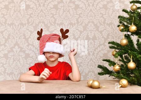 Ein Junge in einer Mütze, in den Augen gekleidet. Ein verwirrter Mann sitzt am Weihnachtsbaum. Stockfoto