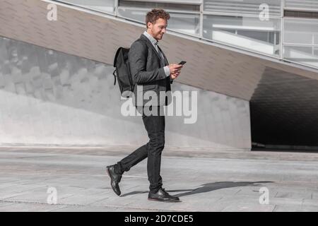 Glücklicher junger Geschäftsmann, der mit dem Telefon unterwegs ist, pendelt zur Arbeit mit einer Rucksack-Tasche auf der Stadtstraße. Geschäftsmann SMS Blick auf Smartphone Stockfoto