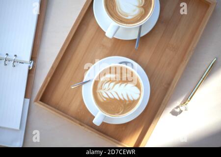 Draufsicht zwei Tassen Kaffee Latte Kunstschaum hält von Frau Hand mit unfokussierten platziert Brillen und Notizblock auf Holztisch. Stockfoto