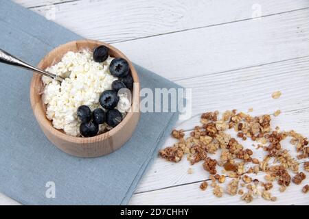 Holzschale mit Bauernkäse, Heidelbeere mit Löffel und Müsli auf blauer Serviette auf weißem Holzhintergrund flach legen. Gesunde Ernährung Konzept. Bildkons Stockfoto