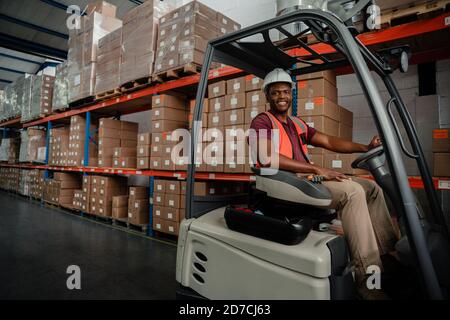 Lächelnder Fabrikarbeiter, der durch das Lager fährt und Pakete für die Lieferung auffüllt. Stockfoto