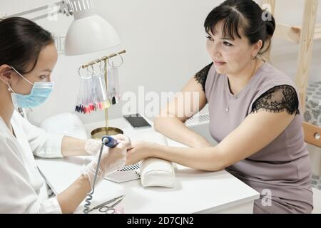 Wunderschöne Frau mittleren Alters bekommen ihre Nägel von einem Manikuristen in einem Schönheitssalon getan. Hardware Maniküre. Echte Menschen. Seitenansicht. Schönheitssalon als kleines Unternehmen. Stockfoto