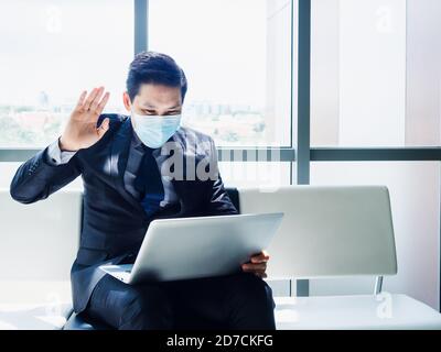 Asiatische Geschäftsmann in Anzug tragen schützende Gesichtsmaske winkte zu Begrüßen Sie die Kollegen auf dem Laptop-Computer-Monitor auf dem Schoß Sitzen im modernen Büro Stockfoto