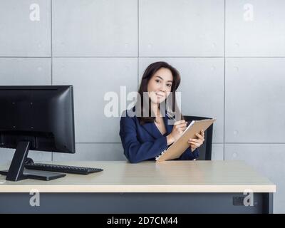 Asiatische Geschäftsfrau lange Haare in blauen Anzug arbeiten mit Desktop-Computer im modernen Büro auf grauen Wand Hintergrund. Schöne Sekretärin hält Stift und Stockfoto