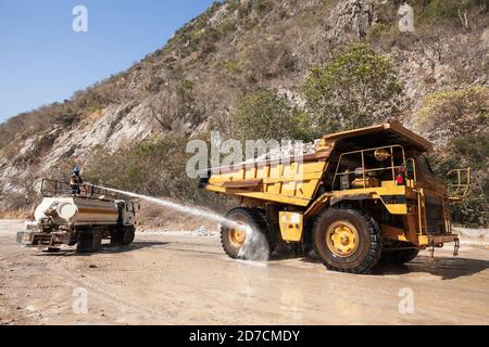 Männlicher Arbeiter auf Wasserwagen sprüht Wasser auf den großen Radwagen in einem Bergbau, Kalksteinberg im Hintergrund. Stockfoto