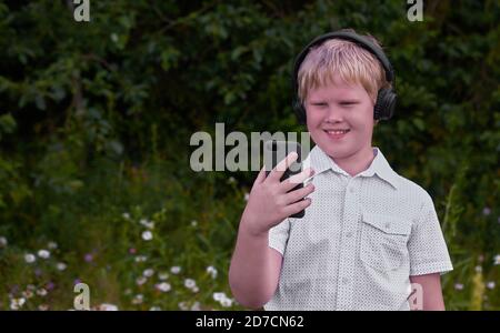 Ein lächelnder Junge mit Kopfhörern und einem Telefon kommt herbei Der Park im Sommer Stockfoto