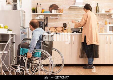 Behinderter Mann im Rollstuhl öffnet den Kühlschrank, während seine Frau kocht. Behinderter gelähmter behinderter Mann mit Gehbehinderung, der sich nach einem Unfall integriert. Stockfoto