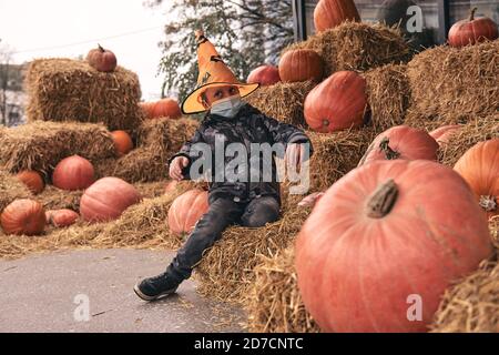 Junge im Halloween Kostüm mit Kürbissen auf dem Bauernmarkt steht auf Heu. Furchterregende Dekorationen. Kinder Trick or Treat. Das Tragen einer schützenden Gesichtsmaske an Halloween Stockfoto