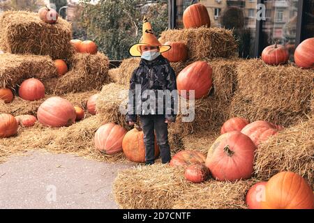 Junge im Halloween Kostüm mit Kürbissen auf dem Bauernmarkt steht auf Heu. Furchterregende Dekorationen. Kinder Trick or Treat. Das Tragen einer schützenden Gesichtsmaske an Halloween Stockfoto